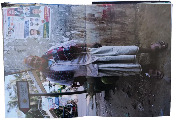 Book spread: an elderly man in a checked lungi and scarf standing against a poster-covered wall, printed sideways across the gutter